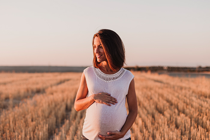 Mulher branca grávida no meio de uma plantação com a mão na barriga, sorrindo e olhando para o lado