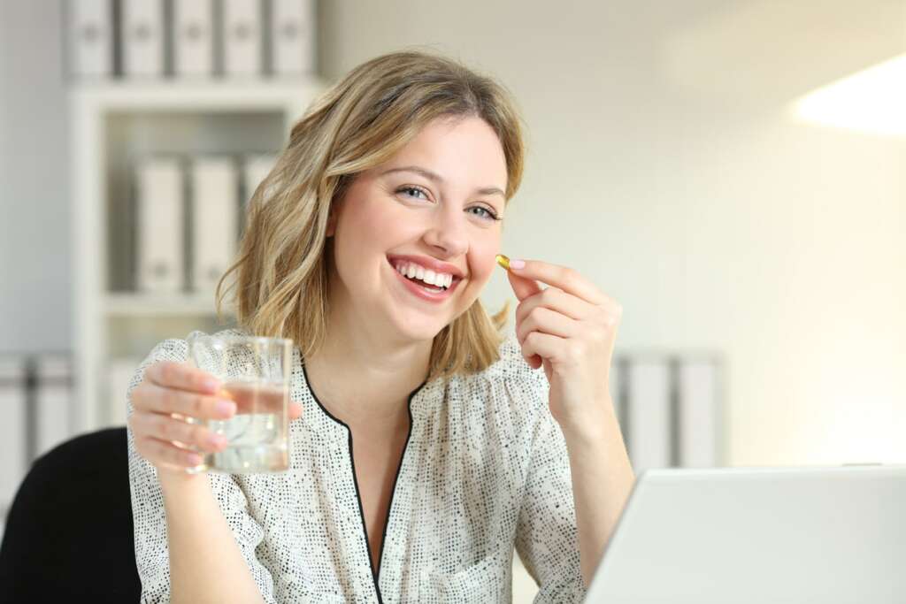 mulher sorridente segurando copo d'agua e um comprimido de colágeno