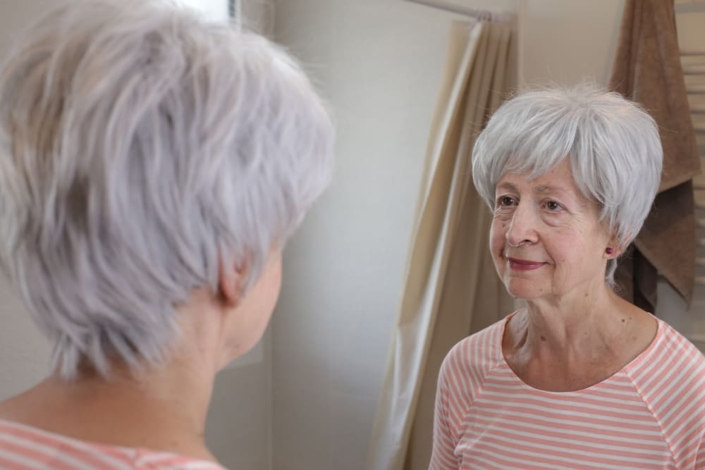 Mulher com cabelos grisalhos se olhando no espelho e notando o envelhecimento da pele