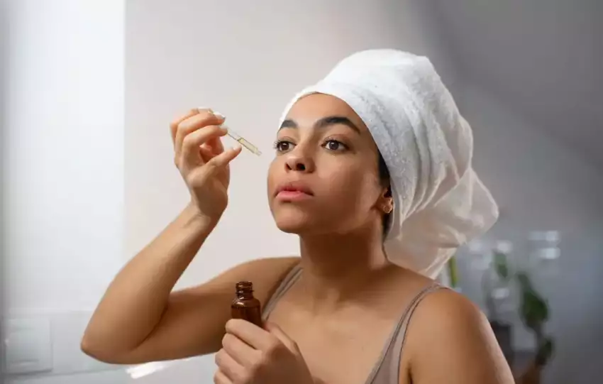 mulher-negra-de-toalha-branca-na-cabeca-passando-serum-facial-em-frente-ao-espelho