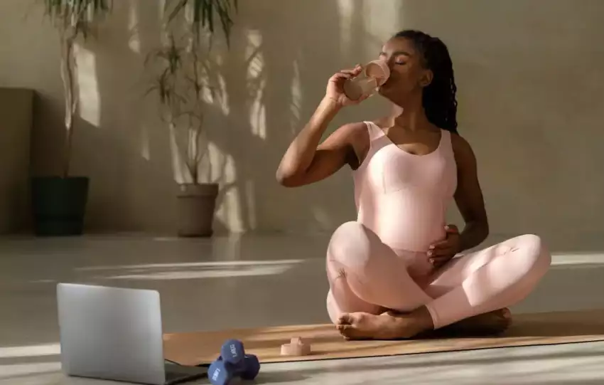 mulher-negra-gravida-usando-roupa-de-academia-rosa-sentada-no-chao-ao-lado-de-um-computador-descansando-depois-de-exercicio-tomando-suplemento